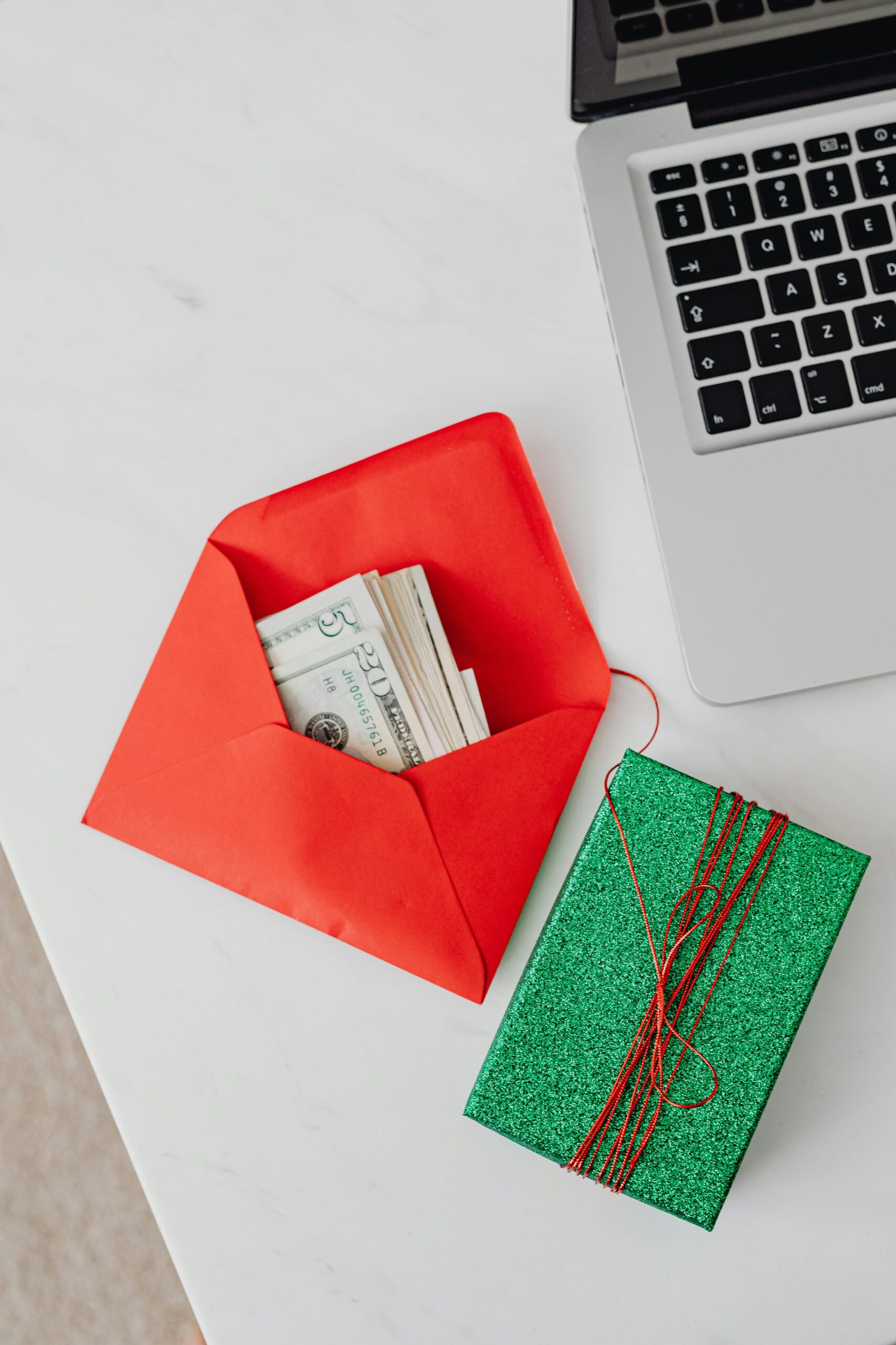 Home A red envelope containing cash beside a laptop and glittering green gift box on a white table.