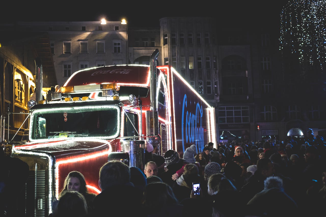 A Coca-Cola branded truck surrounded by a dense crowd during a nighttime parade, illuminated with festive lights.